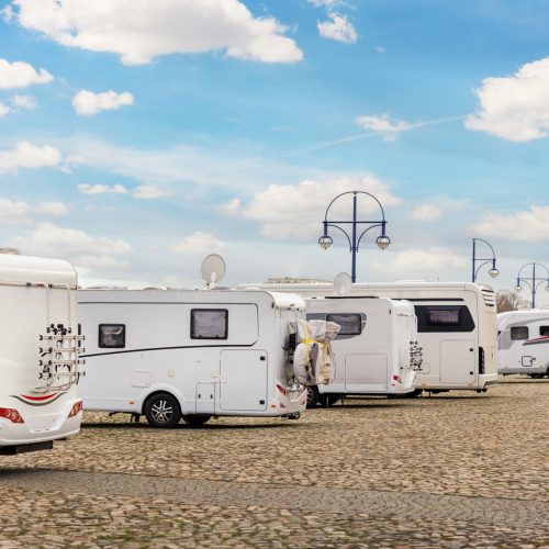 Many white modern campervan recreational motor home vehicles parked in row at camper park site Magdeburg city against Elbe river bridge. Motorhome campground stataion travel destination. RV lifestyle.